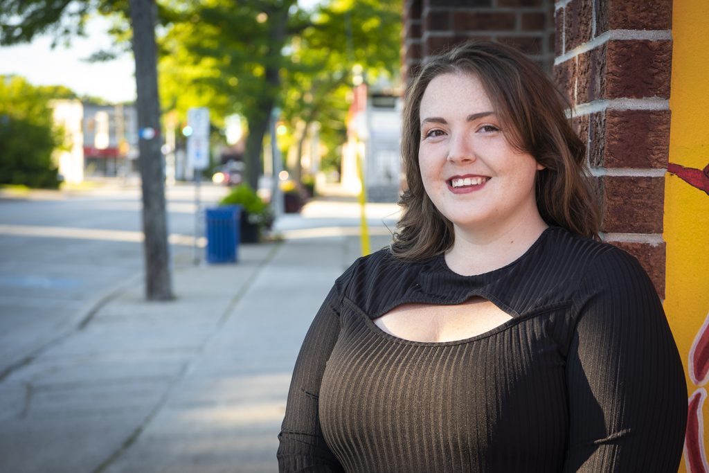 A person with shoulder-length hair and a black shirt on the sidewalk smiling in Mitton Village,