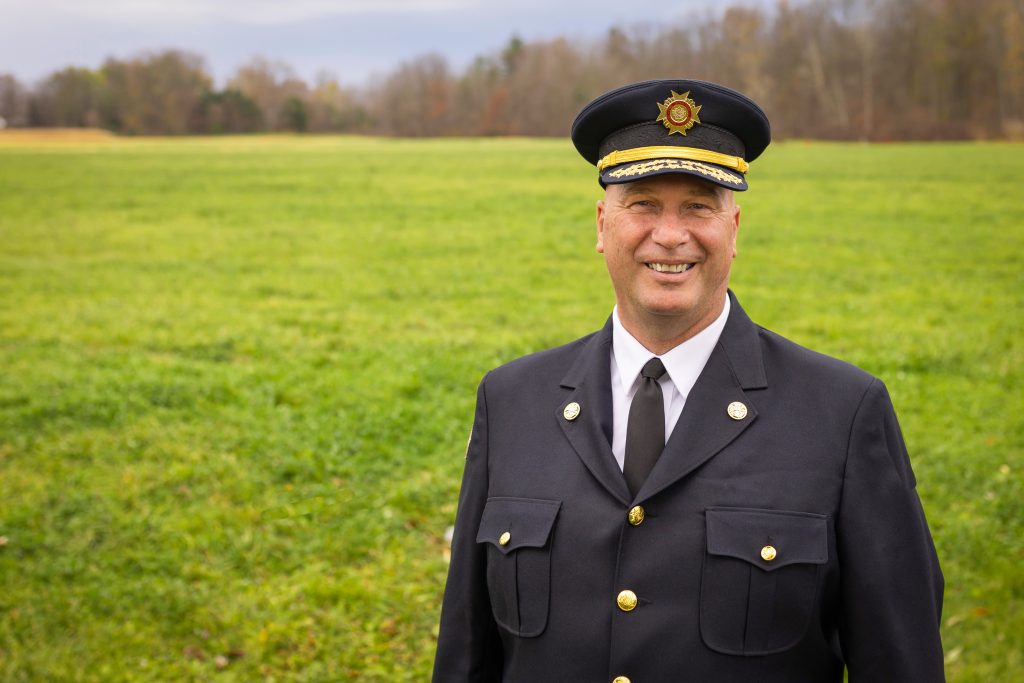 Man wearing fire chief uniform smiling while standing ina  grass field.