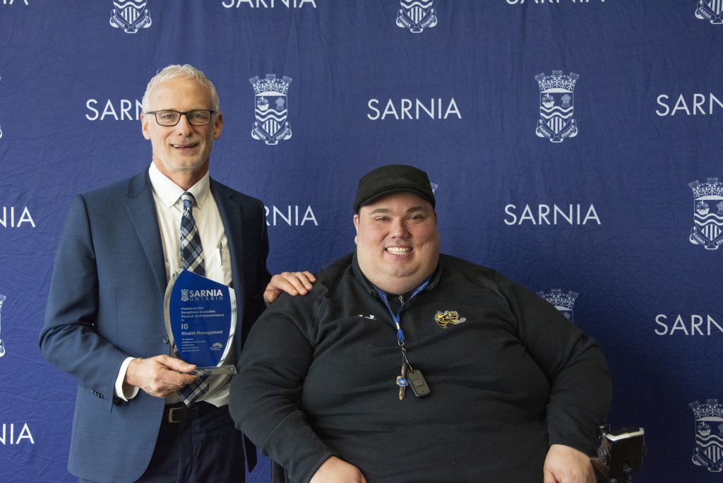 Tow individuals smiling, one holding an award.