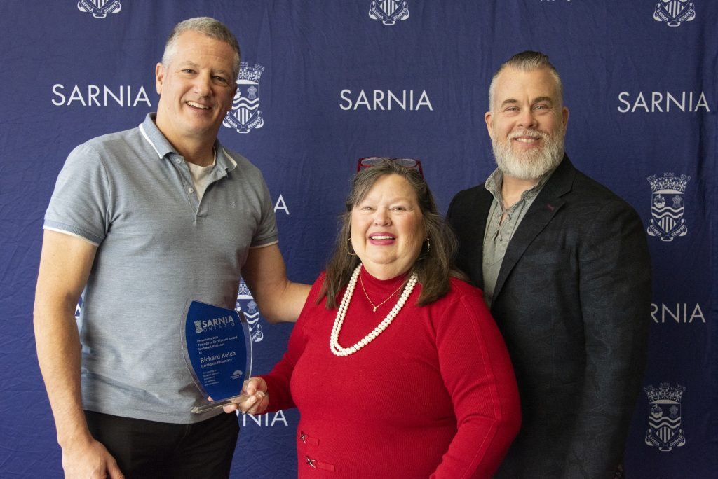 Three individuals smiling, with the one in the centre holding an award.