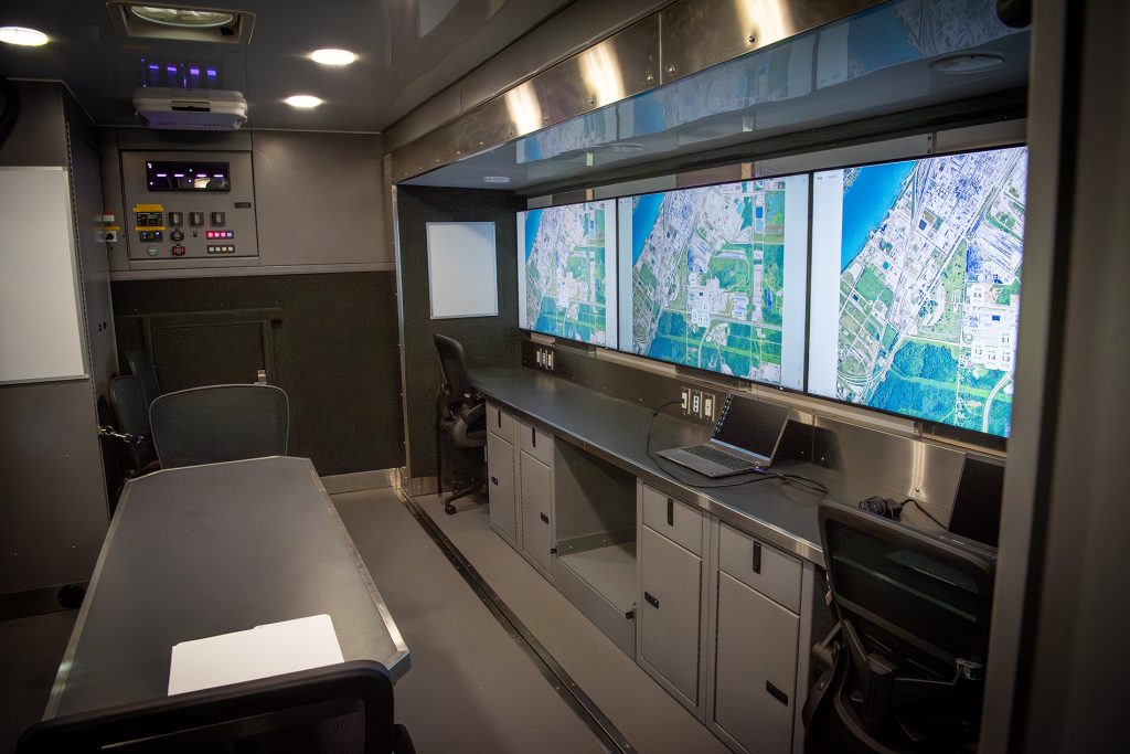 The interior of a mobile command vehicle with several digital workstations featuring large screens.
