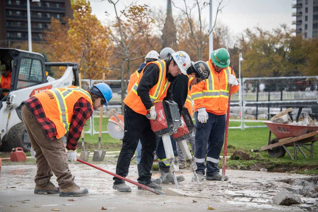 "Construction workers wearing safety vests and helmets are breaking up concrete on a sidewalk using tools, including a jackhammer and shovels. A small loader and wheelbarrow are nearby, with fencing and autumn trees in the background."